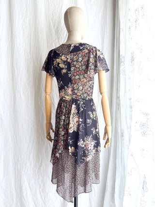 Back view of a vintage 70s bohemian floral dress displayed on a fabric dress form against a white curtain backdrop. The dress features a navy blue base with a multicolor floral pattern.
