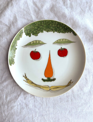 A white ceramic plate with a whimsical vegetable face design, featuring green leafy hair, red tomato eyes, and a carrot nose.