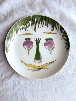 A white ceramic plate with a whimsical hand-painted face made from vegetables. 