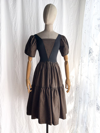 A 1980s vintage dress displayed on a mannequin against a white fabric backdrop. The dress has black bodice  and puff sleeves with a brown and black striped skirt.