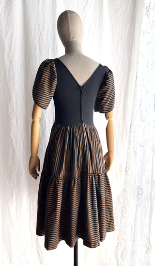 The back view of a 1980s J Lechat dress displayed on a mannequin, featuring a center zipper closure, puffy sleeves and a full striped tiered skirt.