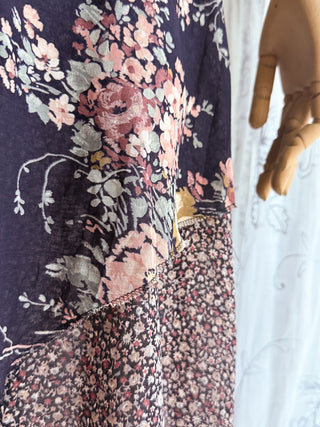 A close-up of a floral dress or garment displayed on a mannequin, featuring a dark navy base with large pink and peach flowers, white and gray leaves, and a contrasting small-scale floral pattern in the lower section.
