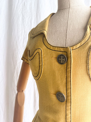 A mustard yellow short-sleeve dress with contrast dark top stitching, a round neckline, and two dark buttons with a cross motif.