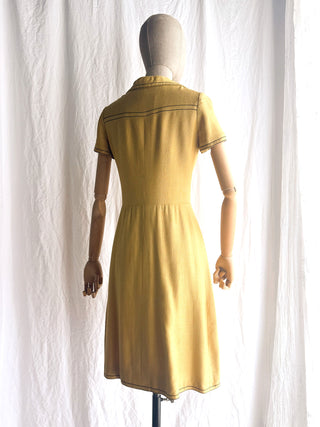 The back view of a mustard yellow vintage French Anne Valone dress from Paris displayed on a mannequin from the back, set against a white fabric backdrop.