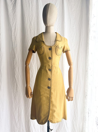 A mustard yellow short-sleeve dress displayed on a wooden mannequin against a white fabric backdrop. The dress features a pointed collar, dark buttons down the front, and two decorative chest pockets.