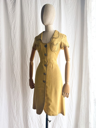The side view of a  vintage French Anne Valone mustard yellow short-sleeve dress displayed on a wooden mannequin against a white curtain backdrop.