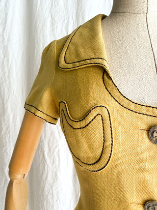 Vintage Anne Valone Paris mustard yellow short-sleeve knit top with black contrast stitching and scalloped edges, displayed on a beige mannequin dress form with a wooden arm against a white fabric backdrop.
