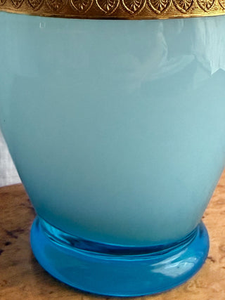 A close-up of a translucent light blue opaline glass vase or container with a shiny blue base and a decorative gold-toned rim, resting on a wooden surface.