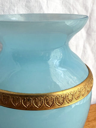 A  vintage blue opaline light blue glass vase with an ornate gold metal band around the neck, shown close up against a white fabric background.