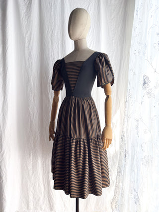 A vintage 70s dress displayed on a mannequin against a white fabric backdrop. The dress has a black bodice with puff sleeves and a square neckline with a tiered skirt in black and brown vertical and horizontal  stripes.