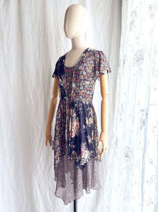 Side view of a vintage 1970s short-sleeve floral dress displayed on a fabric dress form against a white curtain backdrop. The dress features a navy blue base with a multicolor floral pattern.