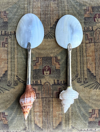 Back view of two vintage spoons with oval mother-of-pearl handles and shell-shaped spoons resting on a patterned fabric background.