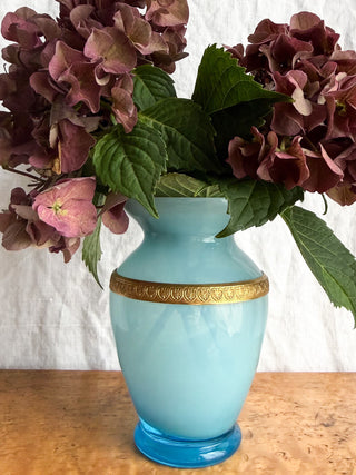 A  French vintage light blue glass vase with a decorative gold band around the neck, filled with dark burgundy hydrangea flowers and green leaves, set on a wooden surface against a white fabric backdrop.