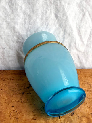 The bottom view of a vintage blue glass vase with a gold decorative band around the neck, resting on a wood surface against a white fabric backdrop.